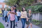 A multi-racial group of university students cheerfully walk down a sidewalk together. One student is carrying his boyfriend on his back.