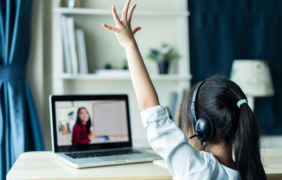A child raising her hand in front of a computer, studying remotely