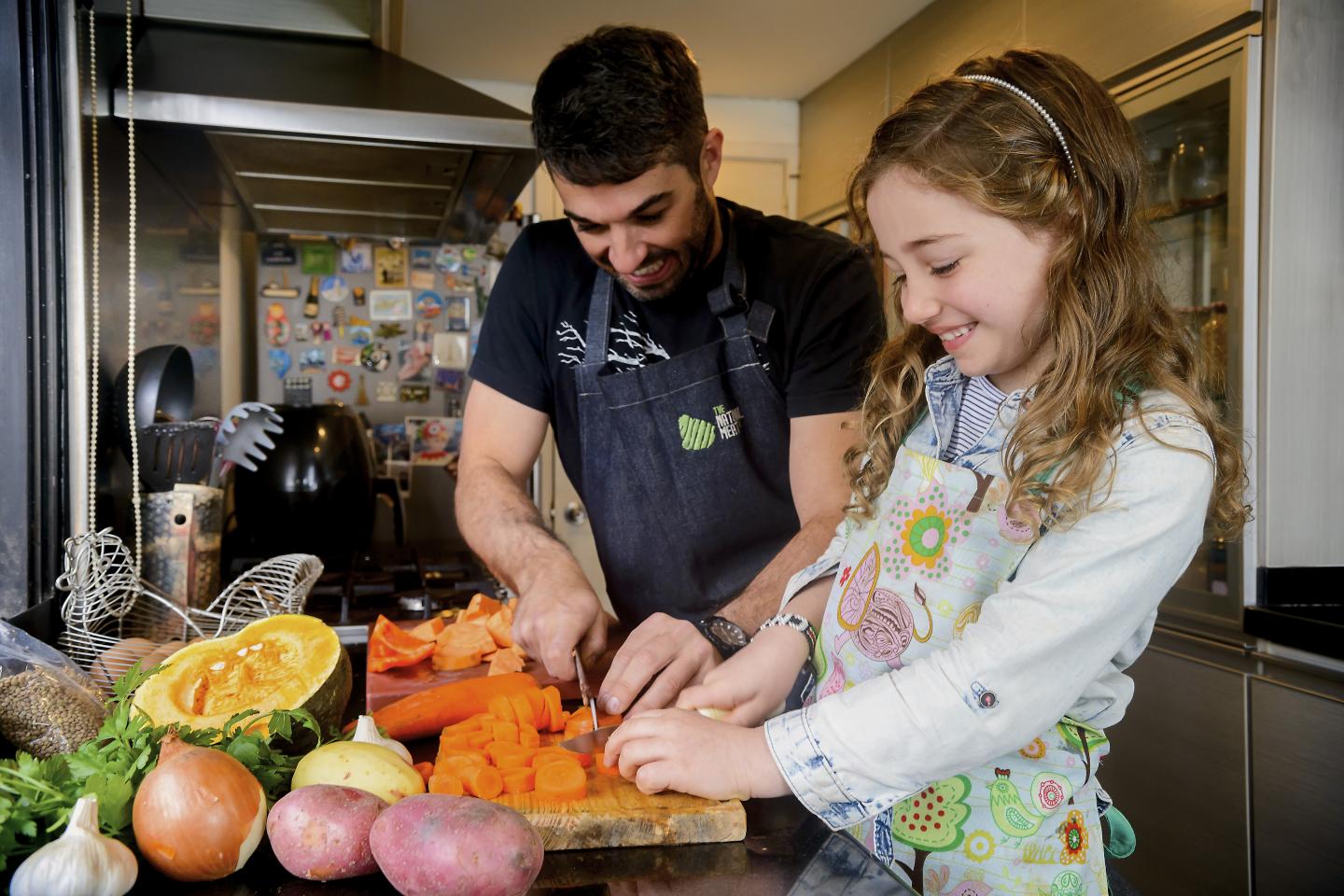 A father cuts carrots while his daughter holds them steady